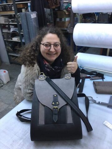 Bu karede atölyemizde eğitim alan katılımcımız, kendi tasarımı ve el emeğiyle ortaya çıkardığı çantasını gururla sergiliyor. Elindeki işaret, memnuniyetin ve başarı hissinin en güzel göstergesi. Arka plandaki üretim ortamı, bu çalışmanın bir parçası olarak sürecin titizlikle yürütüldüğünü yansıtıyor.