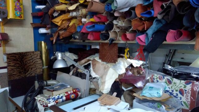 Atölyemizin üretim alanı detaylı bir şekilde görülüyor. Farklı renk ve dokularda rulo halindeki deriler raflarda özenle saklanırken, doğal postlar ve çeşitli aksesuar malzemeleri de üretim için hazır bekliyor. Kesim ve tasarım işlemlerinin yürütüldüğü çalışma masası, üretim sürecinin merkezinde yer alıyor. Her parça, el işçiliği ve özgün tasarımlarla hayat bulmak üzere bu alanda hazırlanıyor.