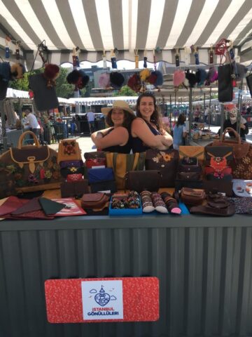 İstanbul Gönüllüleri'nin desteklediği bu etkinlikte, el emeği ürünlerimizin yer aldığı standımız pozitif enerjisiyle dikkat çekiyor. Arkamızdaki çantalar, önde sergilenen cüzdanlar ve rengârenk aksesuarlar tamamen kadın emeğiyle üretilmiş olup, özgün tasarımlarıyla el işçiliğini ön plana çıkarıyor. Güleryüzlü ekip arkadaşlarımız da üretimin ve emeğin gücünü yansıtan bir duruş sergiliyor.