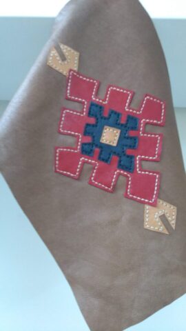 Görselde yer alan bu el işçiliği motif, Selçuklu mimarisinin simetrik ve güçlü çizgilerinden ilham alarak tasarlanmıştır. Kahverengi doğal deri üzerine kontrast renklerle uygulanan bu mozaik desen, tamamen elde kesilmiş ve dikilmiştir. Merkezde yer alan lacivert ve kırmızı kompozisyon, “birlik” ve “denge”yi simgelerken, ok ucu şeklindeki detaylar yön ve korunma anlamı taşır. Estetik olduğu kadar anlamlı da olan bu tasarım, çanta, cüzdan veya aksesuar olarak her ürünün ruhunu zenginleştirir.