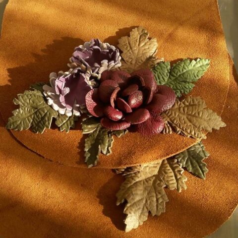 Görselde yer alan çanta kapağı, tamamen el işçiliğiyle hazırlanmış deri çiçek ve yaprak süslemeleriyle dikkat çekiyor. Her bir yaprak ve çiçek detayı özenle kesilip biçimlendirilmiş, derinin doğal dokusu bozulmadan üç boyutlu bir görünüm elde edilmiştir. Zengin toprak tonlarıyla birleşen kırmızı gül ve mor çiçek motifleri, ürüne zarif ve özgün bir karakter kazandırıyor.