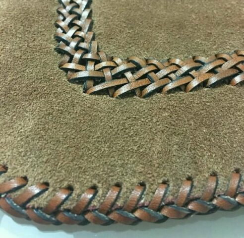 Kahverengi süet deri yüzey üzerine uygulanan örgü teknikli dikiş, hem estetik hem de dayanıklılık açısından öne çıkıyor. Bu tür örgülü kenar işlemleri özellikle çanta, cüzdan ve aksesuar tasarımlarında el emeğini vurgulayan zarif detaylar arasında yer alır. Kullanılan çift renkli deri şeritler, dokuyu daha belirgin ve dinamik hale getiriyor. Titizlikle uygulanmış bu teknik, ustalığın ve sabrın güzel bir göstergesi.